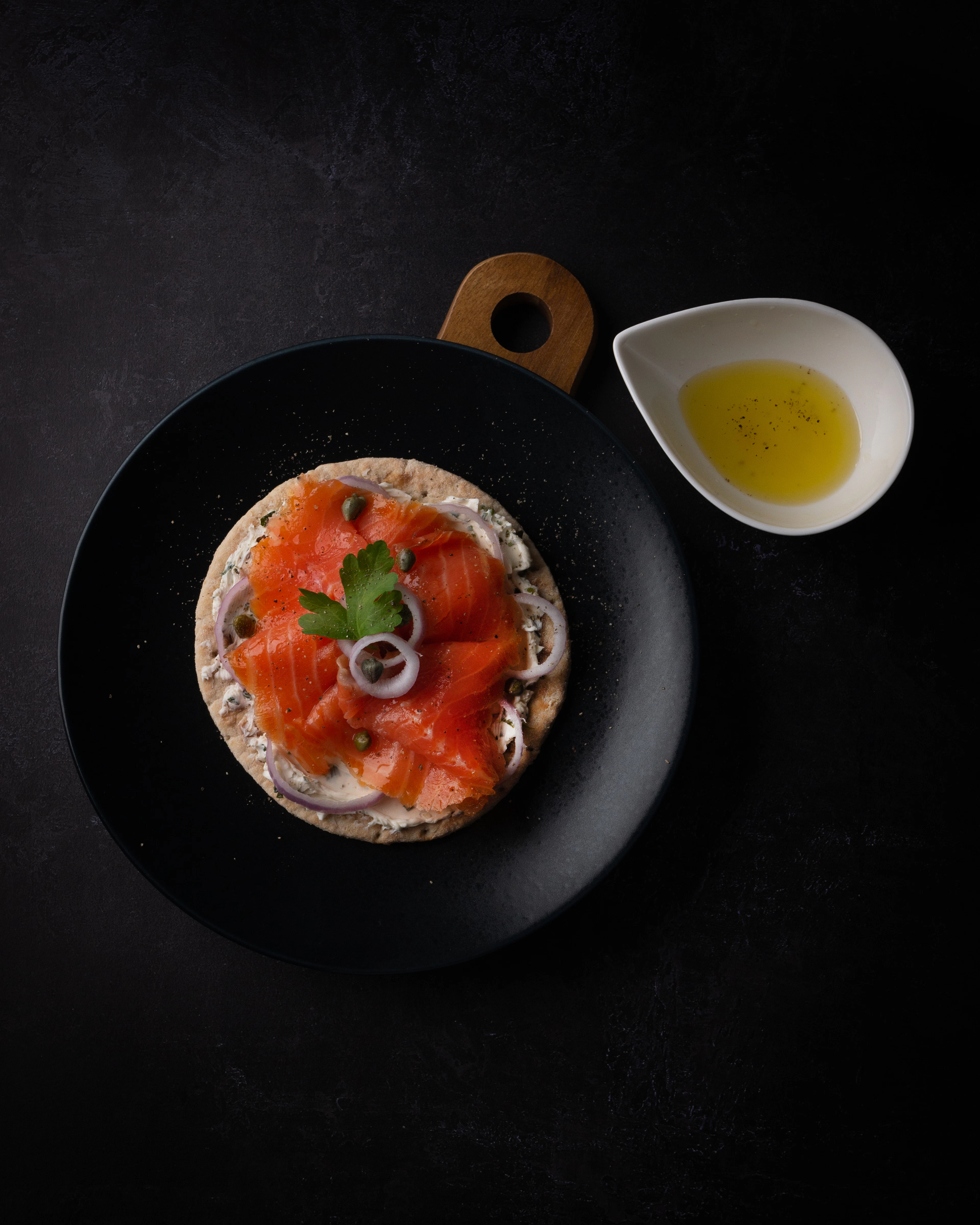 A round Finnish flatbread spread with cream cheese and topped with folded smoked salmon, capers, red onion and dill, plated in Helsinki beside a dish of oil