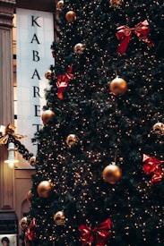 A large Christmas tree decorated with golden baubles and red ribbons is prominently displayed. Bright white lights are wrapped around the tree, creating a festive atmosphere. A vertical sign with the word 'KABARET' is partially visible in the background, and a decorative street lamp can be seen nearby.