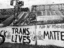 A black and white image displays a wall with various graffiti and murals. The prominent message reads 'TRANS LIVES MATTER' with illustrations of individuals labeled 'Marsha,' 'Tony,' and 'Dominique.' The surface is wet with rain droplets visible on the camera lens, adding a textured effect. Other writings can be seen on the wall, emphasizing messages of empowerment and social justice.