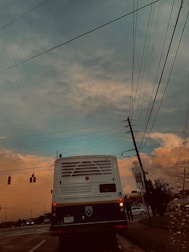 A bus with the Emory University logo is stopped at a traffic light on a road, surrounded by power lines stretching across the sky. The evening sky displays hues of blue and orange, with clouds indicating a possible sunset. A section of the road is visible with a few other vehicles and traffic lights.