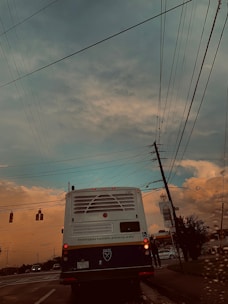 A bus with the Emory University logo is stopped at a traffic light on a road, surrounded by power lines stretching across the sky. The evening sky displays hues of blue and orange, with clouds indicating a possible sunset. A section of the road is visible with a few other vehicles and traffic lights.