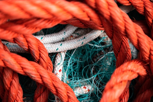 Interwoven ropes of various colors and textures. Thick orange ropes are primarily in the foreground, while white ropes with colored specks and thin blue-green nets are tangled together in the background.