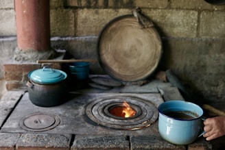 A rustic, traditional kitchen setup features a wood-burning stove with a small fire visible in its circular opening. A person is holding a blue enamel cup with a liquid inside, possibly coffee or tea, near the stove. Several cooking pots with lids, also in blue and black, are resting on the stove. A large wooden lid or plate leans against the stone wall, and a wooden stick or spatula is lying across the pots.