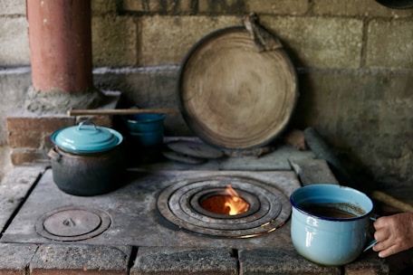 A rustic, traditional kitchen setup features a wood-burning stove with a small fire visible in its circular opening. A person is holding a blue enamel cup with a liquid inside, possibly coffee or tea, near the stove. Several cooking pots with lids, also in blue and black, are resting on the stove. A large wooden lid or plate leans against the stone wall, and a wooden stick or spatula is lying across the pots.