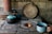 A rustic, traditional kitchen setup features a wood-burning stove with a small fire visible in its circular opening. A person is holding a blue enamel cup with a liquid inside, possibly coffee or tea, near the stove. Several cooking pots with lids, also in blue and black, are resting on the stove. A large wooden lid or plate leans against the stone wall, and a wooden stick or spatula is lying across the pots.