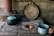 A rustic, traditional kitchen setup features a wood-burning stove with a small fire visible in its circular opening. A person is holding a blue enamel cup with a liquid inside, possibly coffee or tea, near the stove. Several cooking pots with lids, also in blue and black, are resting on the stove. A large wooden lid or plate leans against the stone wall, and a wooden stick or spatula is lying across the pots.
