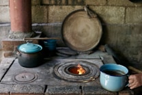 A rustic, traditional kitchen setup features a wood-burning stove with a small fire visible in its circular opening. A person is holding a blue enamel cup with a liquid inside, possibly coffee or tea, near the stove. Several cooking pots with lids, also in blue and black, are resting on the stove. A large wooden lid or plate leans against the stone wall, and a wooden stick or spatula is lying across the pots.