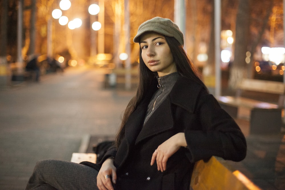 Frau in schwarzem Mantel und braunem Hut sitzt nachts auf der Bank