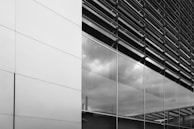 A modern building facade featuring large reflective glass panels and sleek metal cladding. The glass reflects cloudy skies and industrial structures, creating a clean and contemporary look.