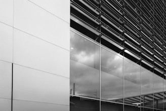 A modern building facade featuring large reflective glass panels and sleek metal cladding. The glass reflects cloudy skies and industrial structures, creating a clean and contemporary look.