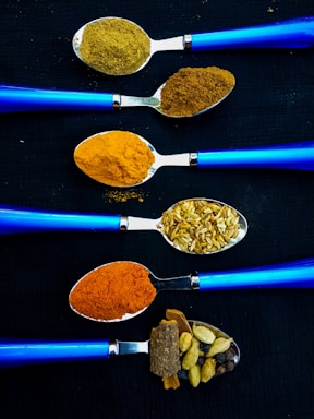 Five blue-handled spoons arranged horizontally on a dark background. Each spoon contains different spices and ingredients: ground coriander, ground cumin, turmeric, fennel seeds, red chili powder, and whole spices like cinnamon and cardamom. The vibrant colors of the spices contrast against the sleek blue of the spoons.
