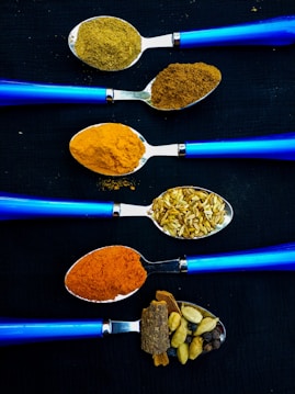 Five blue-handled spoons arranged horizontally on a dark background. Each spoon contains different spices and ingredients: ground coriander, ground cumin, turmeric, fennel seeds, red chili powder, and whole spices like cinnamon and cardamom. The vibrant colors of the spices contrast against the sleek blue of the spoons.