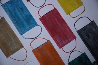 A collection of various colored face masks laid out on a flat surface. The masks include shades of blue, red, yellow, black, and orange, each with matching or contrasting ear loops. They are arranged in a scattered pattern, creating a colorful and diverse display.
