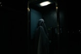 A person dressed as a ghost with a sheet over their head, standing inside a dimly lit telephone booth. The atmosphere is eerie, with minimal lighting highlighting the ghostly figure.