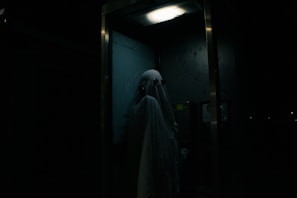A person dressed as a ghost with a sheet over their head, standing inside a dimly lit telephone booth. The atmosphere is eerie, with minimal lighting highlighting the ghostly figure.