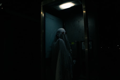A person dressed as a ghost with a sheet over their head, standing inside a dimly lit telephone booth. The atmosphere is eerie, with minimal lighting highlighting the ghostly figure.