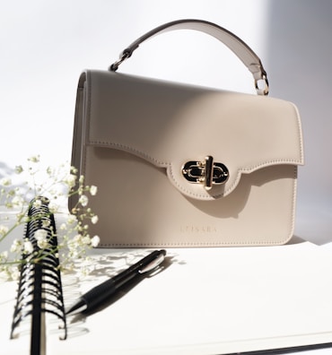 A beige leather handbag with a gold clasp sits on a white surface. To the left, there is a spiral notebook with a pen resting on it. Delicate white flowers are subtly visible, adding a touch of nature to the scene.