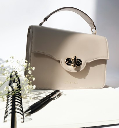 A beige leather handbag with a gold clasp sits on a white surface. To the left, there is a spiral notebook with a pen resting on it. Delicate white flowers are subtly visible, adding a touch of nature to the scene.