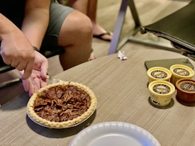 A hand holding a metal pie server is positioned over a pecan pie. The pie rests on a wooden table beside three small containers of ice cream and a smartphone. The background includes the lower legs of a person seated on a black chair.