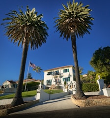 A large white mansion is framed by two tall palm trees. The house has a stepped entrance with a gated driveway and neatly maintained lawns. An American flag flutters on a flagpole to the left of the driveway. The sky is clear and blue, providing a bright backdrop to the scene.