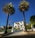 A large white mansion is framed by two tall palm trees. The house has a stepped entrance with a gated driveway and neatly maintained lawns. An American flag flutters on a flagpole to the left of the driveway. The sky is clear and blue, providing a bright backdrop to the scene.