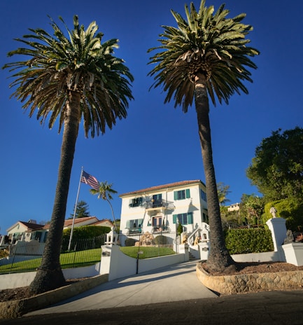 A large white mansion is framed by two tall palm trees. The house has a stepped entrance with a gated driveway and neatly maintained lawns. An American flag flutters on a flagpole to the left of the driveway. The sky is clear and blue, providing a bright backdrop to the scene.