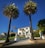 A large white mansion is framed by two tall palm trees. The house has a stepped entrance with a gated driveway and neatly maintained lawns. An American flag flutters on a flagpole to the left of the driveway. The sky is clear and blue, providing a bright backdrop to the scene.