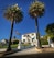 A large white mansion is framed by two tall palm trees. The house has a stepped entrance with a gated driveway and neatly maintained lawns. An American flag flutters on a flagpole to the left of the driveway. The sky is clear and blue, providing a bright backdrop to the scene.