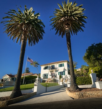 A large white mansion is framed by two tall palm trees. The house has a stepped entrance with a gated driveway and neatly maintained lawns. An American flag flutters on a flagpole to the left of the driveway. The sky is clear and blue, providing a bright backdrop to the scene.