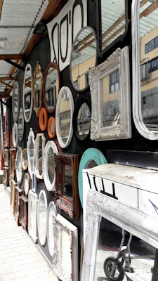 A variety of decorative mirrors with ornate frames are displayed on a wall. The frames are of different shapes and sizes including oval, round, and rectangular. There are also different colors and materials, such as white, turquoise, gold, and wood. The mirrors reflect parts of a building visible in the background. A partially visible wheeled cart is on the ground near a large white-framed mirror.