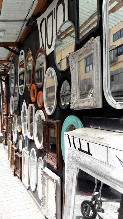 A variety of decorative mirrors with ornate frames are displayed on a wall. The frames are of different shapes and sizes including oval, round, and rectangular. There are also different colors and materials, such as white, turquoise, gold, and wood. The mirrors reflect parts of a building visible in the background. A partially visible wheeled cart is on the ground near a large white-framed mirror.