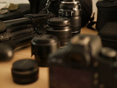 A collection of photography equipment is spread on a surface, including several camera lenses, a camera, a tripod, and camera bags. The focus is on the lenses, with some objects in the background appearing blurred due to the shallow depth of field.