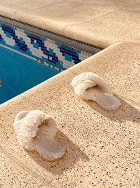 A pair of fluffy white slippers is placed on a textured, beige poolside surface. The surface is adjacent to a pool, visible on the left, with blue and white mosaic tiles creating a decorative border.
