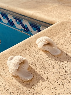 A pair of fluffy white slippers is placed on a textured, beige poolside surface. The surface is adjacent to a pool, visible on the left, with blue and white mosaic tiles creating a decorative border.