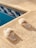 A pair of fluffy white slippers is placed on a textured, beige poolside surface. The surface is adjacent to a pool, visible on the left, with blue and white mosaic tiles creating a decorative border.