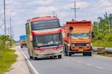A colorful bus and an orange truck are traveling along a rural road with greenery on either side and a cloudy sky above. The bus has distinctive red and white markings and is labeled with 'Patas VIP' and 'Bintang Utara', while the truck is loaded and covered with a protective sheet.