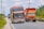 A colorful bus and an orange truck are traveling along a rural road with greenery on either side and a cloudy sky above. The bus has distinctive red and white markings and is labeled with 'Patas VIP' and 'Bintang Utara', while the truck is loaded and covered with a protective sheet.