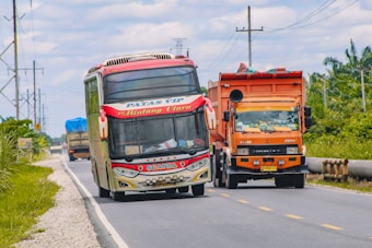 A colorful bus and an orange truck are traveling along a rural road with greenery on either side and a cloudy sky above. The bus has distinctive red and white markings and is labeled with 'Patas VIP' and 'Bintang Utara', while the truck is loaded and covered with a protective sheet.