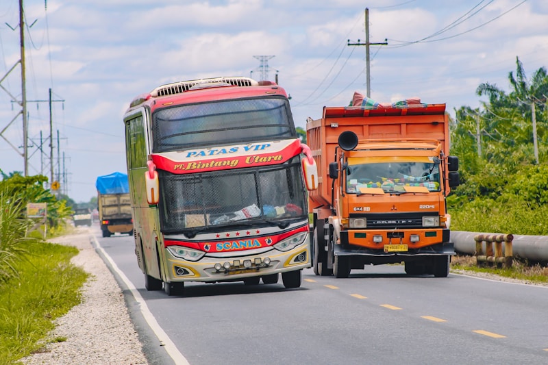 A colorful bus and an orange truck are traveling along a rural road with greenery on either side and a cloudy sky above. The bus has distinctive red and white markings and is labeled with 'Patas VIP' and 'Bintang Utara', while the truck is loaded and covered with a protective sheet.