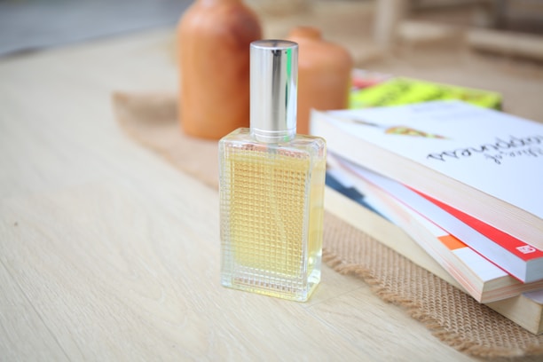 A clear glass perfume bottle with a silver cap is placed on a light wooden surface. Beside it, there are stacked books with colorful covers, and a piece of burlap. In the background, two terracotta-colored ceramic vases add a touch of warmth and earthy tones.
