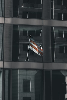 A distortion of a multi-story glass building reflects another structure, with a flag featuring red and white stripes prominently displayed in one of the windows. The building has a grid of reflective, tinted glass panels.