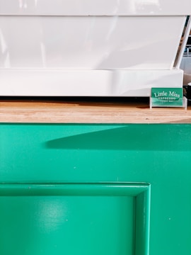 A minimalistic setup featuring a white countertop surface and a wooden edge. Below is a vibrant green panel, likely part of a counter or kiosk. A small green placard with text sits near the corner of the white surface.