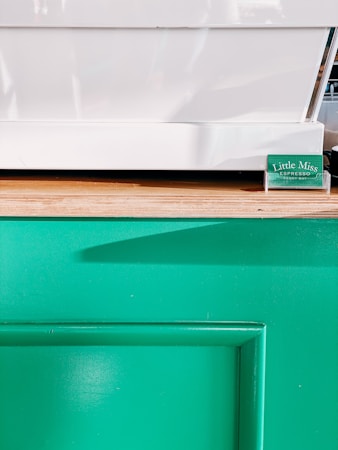 A minimalistic setup featuring a white countertop surface and a wooden edge. Below is a vibrant green panel, likely part of a counter or kiosk. A small green placard with text sits near the corner of the white surface.