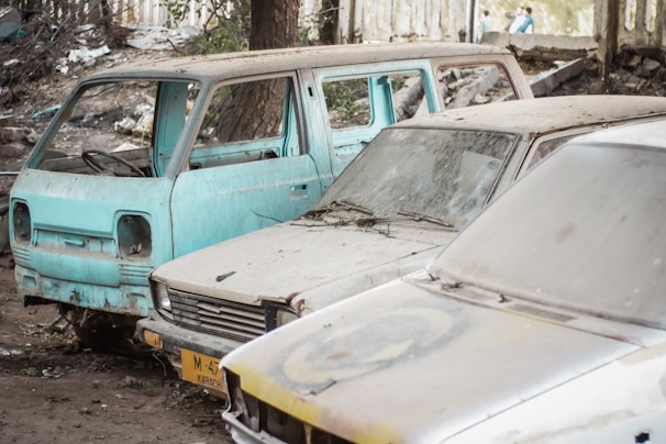 Three abandoned cars are parked in a rundown outdoor area. The vehicles appear old and are covered in dirt and rust, with windows either broken or missing. Surrounding the cars is a littered and untidy environment with debris scattered on the ground. A tree trunk is partially visible behind the vehicles, and there is a wall in the background.