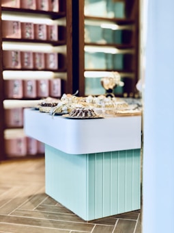 A light turquoise table displaying an array of beautifully arranged desserts, possibly cakes or pastries, with decorative ribbons. In the background, wooden shelves are stocked with rows of pink and blue packages, creating a visually appealing and organized setting.