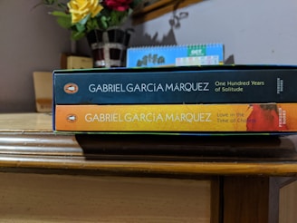 Two books by Gabriel García Márquez are stacked on a wooden surface. The top book is 'One Hundred Years of Solitude' with a dark blue spine, and the bottom book is 'Love in the Time of Cholera' featuring a yellow and orange spine. A vase with red and yellow flowers and a calendar are in the background.