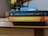 Two books by Gabriel García Márquez are stacked on a wooden surface. The top book is 'One Hundred Years of Solitude' with a dark blue spine, and the bottom book is 'Love in the Time of Cholera' featuring a yellow and orange spine. A vase with red and yellow flowers and a calendar are in the background.