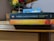 Two books by Gabriel García Márquez are stacked on a wooden surface. The top book is 'One Hundred Years of Solitude' with a dark blue spine, and the bottom book is 'Love in the Time of Cholera' featuring a yellow and orange spine. A vase with red and yellow flowers and a calendar are in the background.