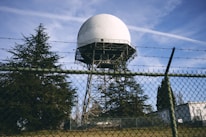 A large radar dome is situated on a metal framework, surrounded by a chain-link fence topped with barbed wire. Evergreen trees are visible in the foreground and background, and the sky is clear with contrails.