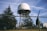 A large radar dome is situated on a metal framework, surrounded by a chain-link fence topped with barbed wire. Evergreen trees are visible in the foreground and background, and the sky is clear with contrails.
